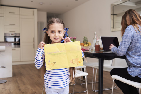 Portrait of smiling little girl showing drawing while her mother working on laptop in the background stock photo