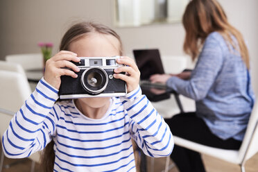 Kleines Mädchen fotografiert mit der Kamera, während ihre Mutter im Hintergrund am Laptop arbeitet - IGGF00017