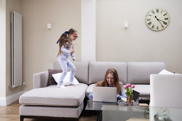 Kleines Mädchen mit Sonnenbrille springt auf dem Sofa, während ihre Mutter am Couchtisch am Laptop arbeitet - IGGF00018