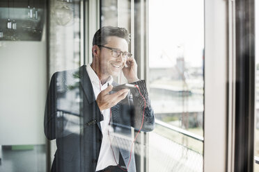 Porträt eines zuversichtlichen Geschäftsmannes am Telefon, der durch ein Fenster schaut - UUF11219