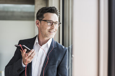 Portrait of businessman standing speaking notes on smartphone while looking through window - UUF11220