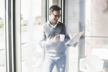 Smiling businessman with coffee mug looking at documents in an office - UUF11242