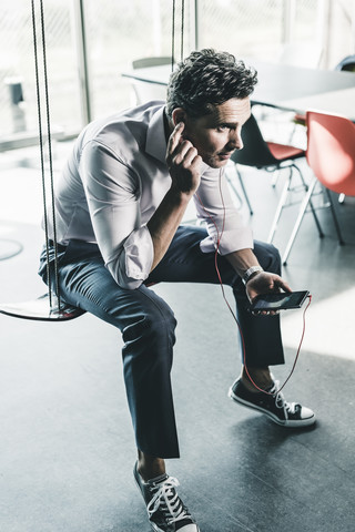 Geschäftsmann im Büro, der auf einer Schaukel sitzt und ein Smartphone benutzt, lizenzfreies Stockfoto
