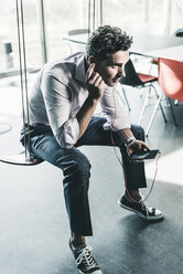 Businessman in office sitting on swing, using smartphone - UUF11255