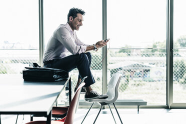 Businessman in office sitting on desk, using smartphone - UUF11257