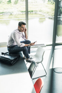 Geschäftsmann im Büro, der auf dem Schreibtisch sitzt und ein Smartphone benutzt - UUF11259