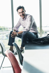 Businessman in office sitting on desk, smiling - UUF11260