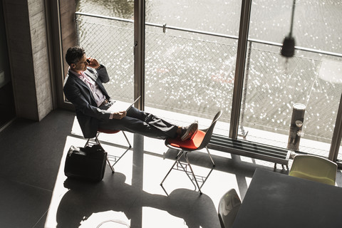 Geschäftsmann, der mit hochgelegten Füßen am Fenster sitzt, am Laptop arbeitet und einen Anruf tätigt, lizenzfreies Stockfoto