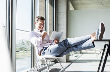 Businessman sitting at desk with feet up, using digital tablet - UUF11285