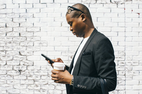 Mittlerer erwachsener Mann vor weißer Backsteinmauer, hält Smartphone und Tasse Kaffee - JUBF00241