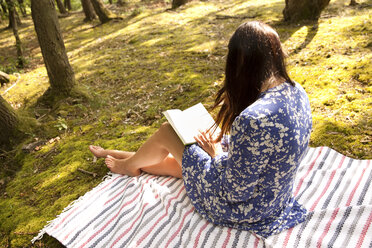 Junge Frau sitzt auf einer Decke im Wald und liest ein Buch - MFRF00940