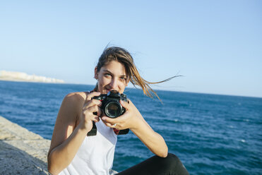 Portrait of smiling young woman with camera at the sea - KIJF01673