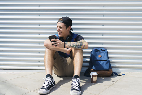 Smiling young man with smartphone, backpack and coffee to go sitting on the ground stock photo