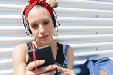 Porträt einer jungen Frau mit Kopfhörern, die auf ihr Smartphone schaut - FMOF00298