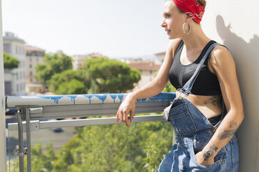Tätowierte junge Frau steht auf einem Balkon und schaut in die Ferne - FMOF00311