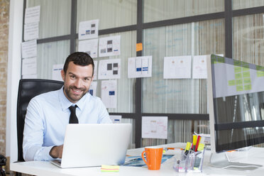 Porträt eines lächelnden Geschäftsmannes im Büro mit Laptop am Schreibtisch - FKF02484