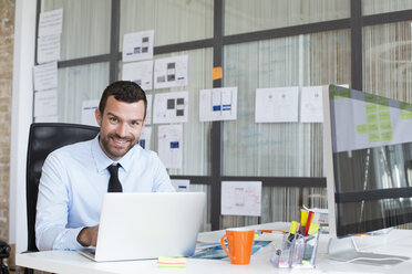 Portrait of smiling businessman in office using laptop at desk - FKF02484