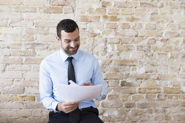 Smiling businessman reading document at brick wall - FKF02505