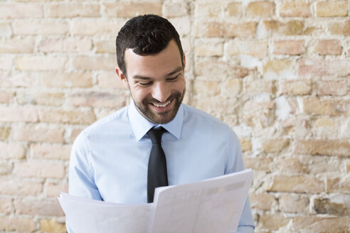 Smiling businessman reading document at brick wall - FKF02506