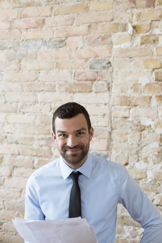 Porträt eines lächelnden Geschäftsmannes mit Dokumenten an einer Backsteinmauer, lizenzfreies Stockfoto