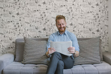 Smiling businessman reading newspaper on couch in his living room - MOMF00201