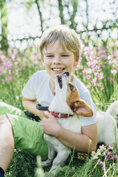 Porträt eines lächelnden kleinen Jungen, der mit seinem Hund auf einer Wiese im Garten sitzt - MJF02132