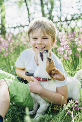 Portrait of smiling little boy sitting with his dog on meadow in the garden - MJF02132