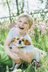 Portrait of laughing little boy sitting with his dog on meadow in the garden - MJF02133