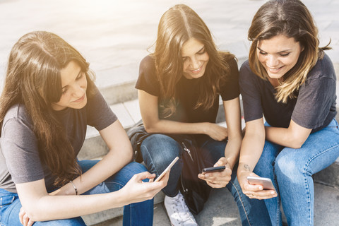 Three smiling young women sitting outdoors looking at cell phones stock photo
