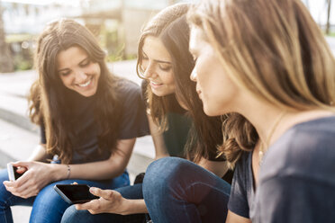 Three happy young women sitting outdoors looking at cell phone - GIOF02987