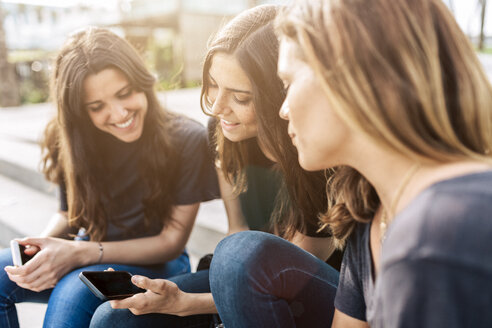 Drei glückliche junge Frauen sitzen im Freien und schauen auf ihr Mobiltelefon - GIOF02987