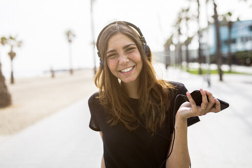 Porträt einer glücklichen jungen Frau an der Strandpromenade, die Musik hört - GIOF02991