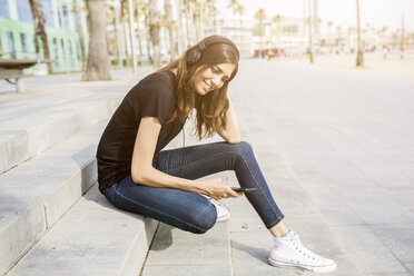 Lächelnde junge Frau sitzt auf einer Treppe und hört Musik - GIOF02992