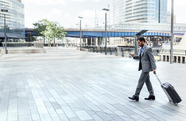 Businessman walking through the city with rolling suitcase and cell phone - MGOF03485