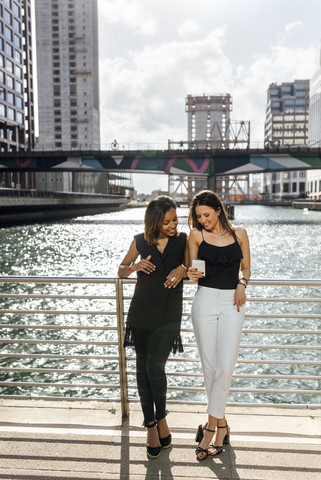 Zwei Frauen auf einer Brücke, die sich in der Stadt ein Mobiltelefon teilen, lizenzfreies Stockfoto