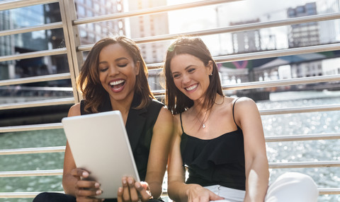 Zwei glückliche Frauen sitzen auf einer Brücke und teilen sich ein Tablet in der Stadt, lizenzfreies Stockfoto