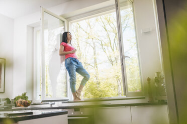 Smiling woman standing in kitchen on windowsill - JOSF01248
