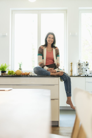 Porträt einer lächelnden Frau, die zu Hause auf dem Küchentisch sitzt, lizenzfreies Stockfoto