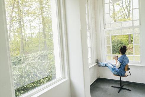 Frau mit einer Tasse Kaffee auf einem Stuhl am Fenster sitzend - JOSF01299