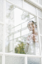 Lächelnde Frau mit einer Tasse Kaffee, die aus dem Fenster schaut - JOSF01308