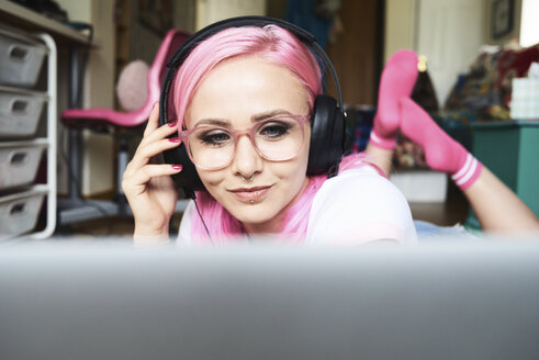 Junge Frau mit rosa Haaren, die zu Hause über einen Laptop Musik hört - IGGF00060