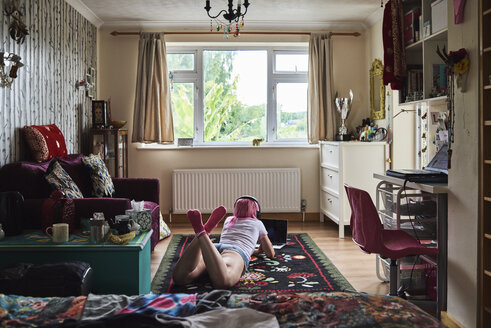 Young woman with pink hair lying on carpet wearing headphones and using laptop at home - IGGF00061