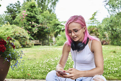 Junge Frau mit rosa Haaren trägt Kopfhörer und benutzt ein Handy im Garten - IGGF00073