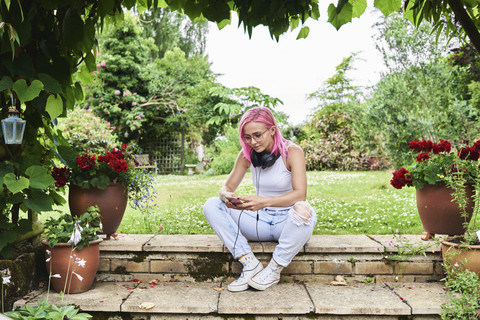 Junge Frau mit rosa Haaren trägt Kopfhörer und benutzt ein Handy im Garten, lizenzfreies Stockfoto