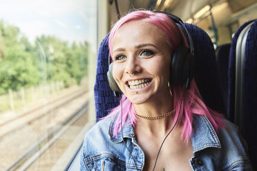 Glückliche junge Frau mit rosa Haaren, die während einer Zugfahrt Musik hört - IGGF00081