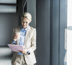 Businesswoman with coffee reading document at the window - UUF11388