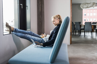 Businesswoman sitting on couch with feet up using tablet - UUF11422