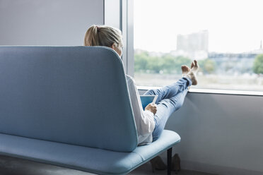 Frau sitzt mit hochgelegten Füßen am Fenster und benutzt ein Tablet - UUF11427