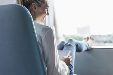 Smiling woman sitting by the window with feet up using tablet - UUF11428