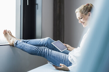 Lächelnde Frau, die mit hochgelegten Füßen am Fenster sitzt und Tablet und Mobiltelefon benutzt - UUF11429